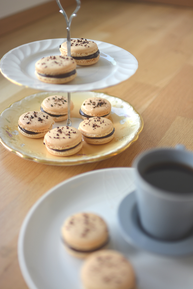 two-tiered cake plate elegantly arranged with chocolate coffee macarons, showcasing their smooth, glossy shells, two additional chocolate macarons sit beside the plate, accompanied by a steaming cup of coffee
