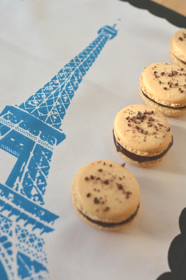 half view of four chocolate coffee macarons arranged in a row on a tea towel, each macaron is garnished with generous chocolate chunks