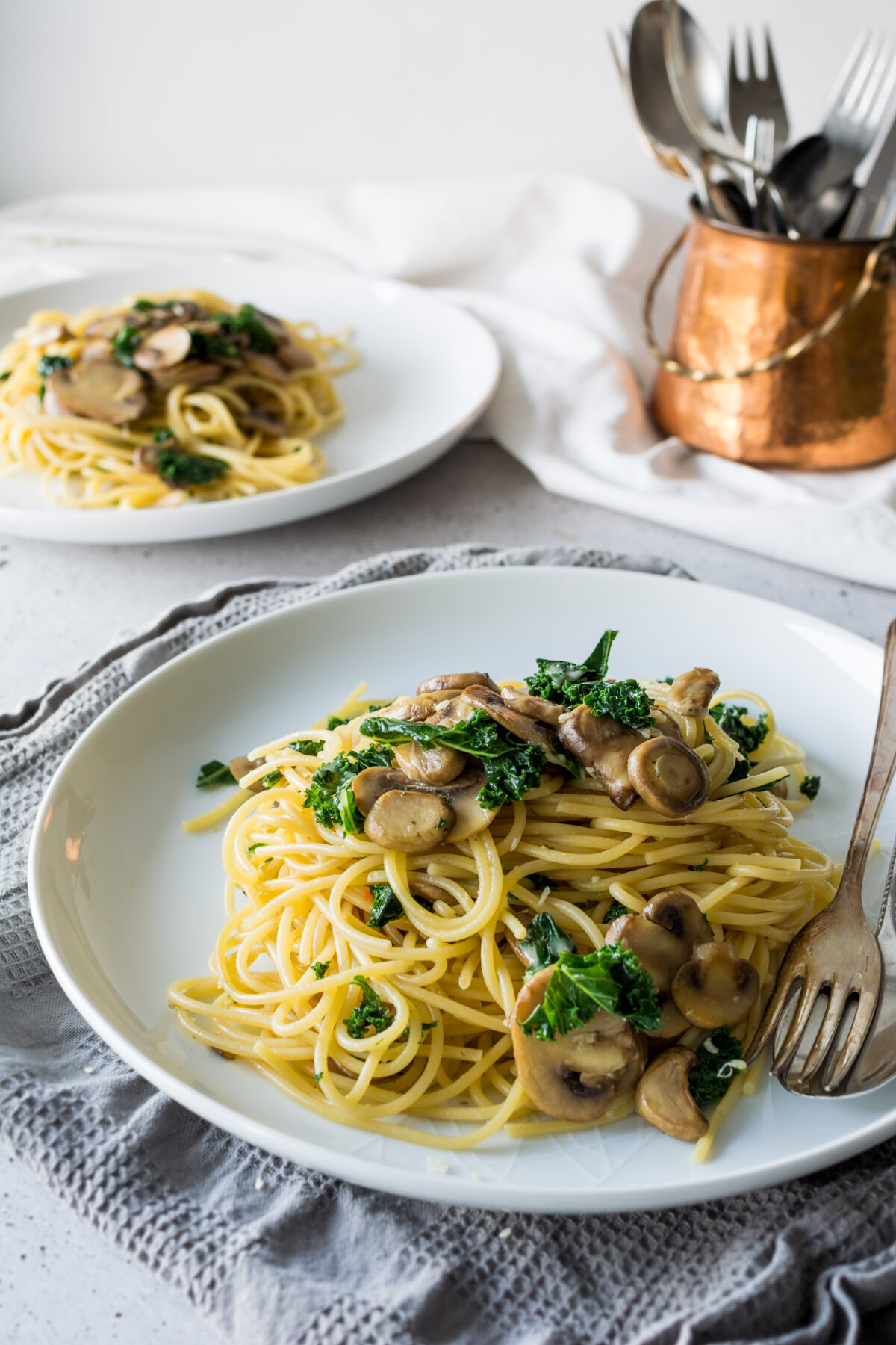 Mushroom, kale, and garlic pasta on a white plate with another plate of the meal in the background and a copper pot with silverware.
