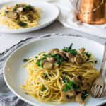 Mushroom, kale, and garlic pasta on a white plate with another plate of the meal in the background and a copper pot with silverware.