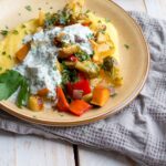 a cropped image of a colorful roasted vegetables, creamy polenta, and zesty herby yogurt sauce on a plate