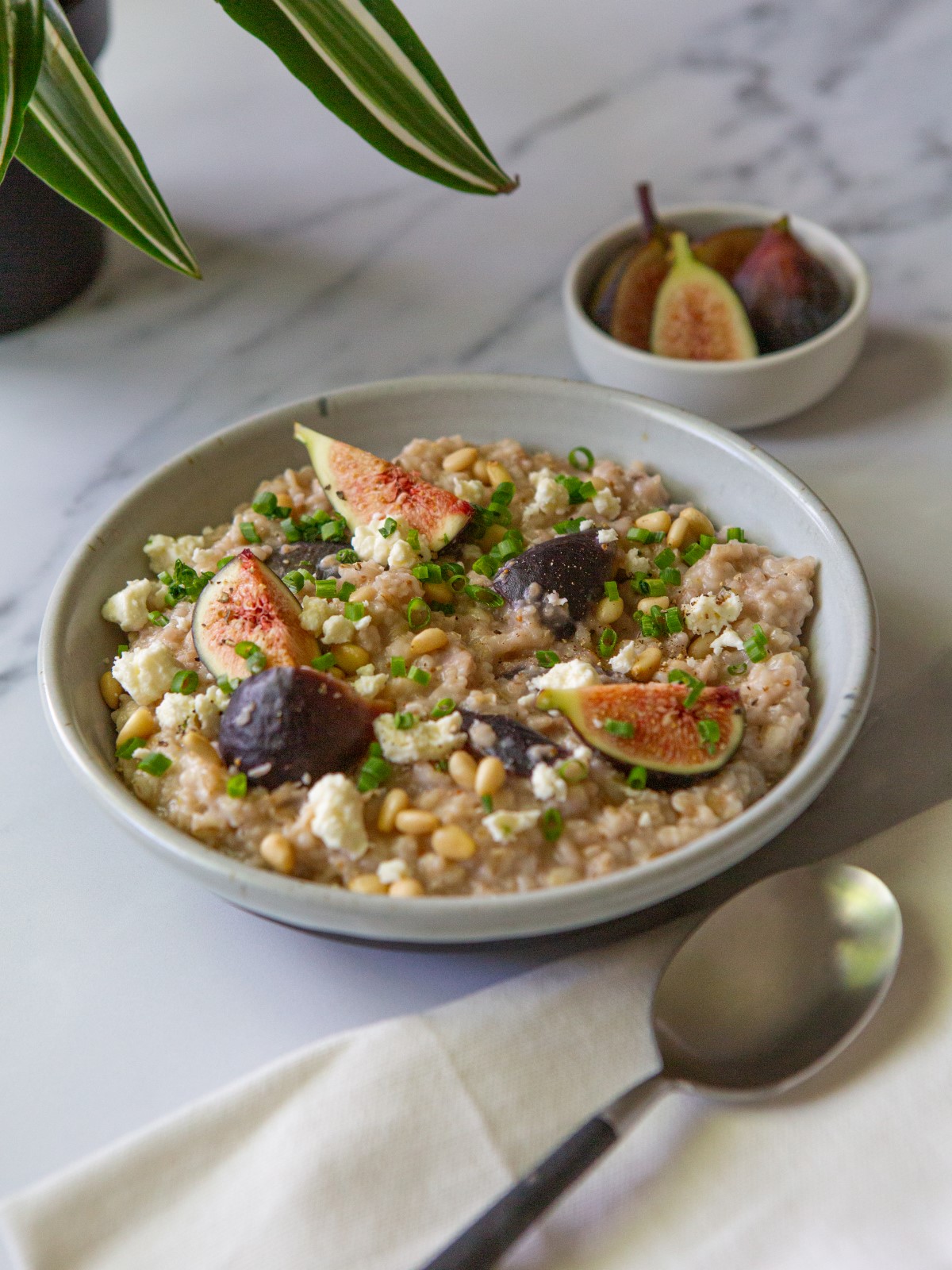 A white bowl filled with savory oatmeal with figs and feta and a napkin with a spoon with a small bowl filled with figs.