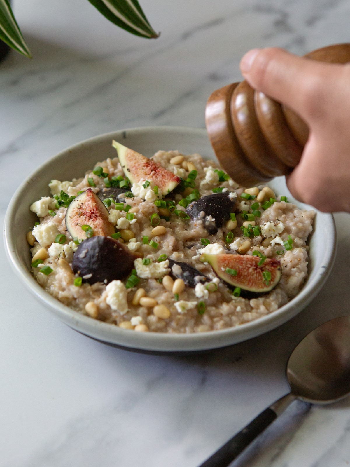 A white bowl filled with savory oatmeal with figs and feta and a spoon. A hand is holding a black pepper grinder.